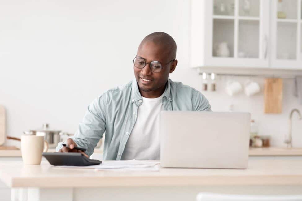 Man using a calculator and laptop at a kitchen counter while reviewing financial documents