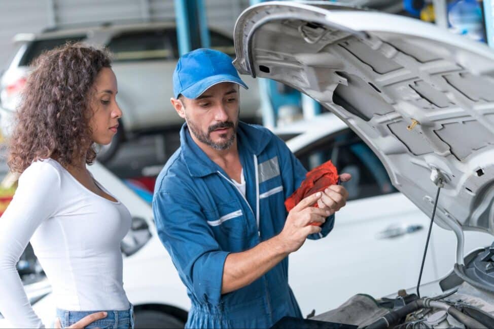 A mechanic in blue coveralls shows a car engine to a customer
