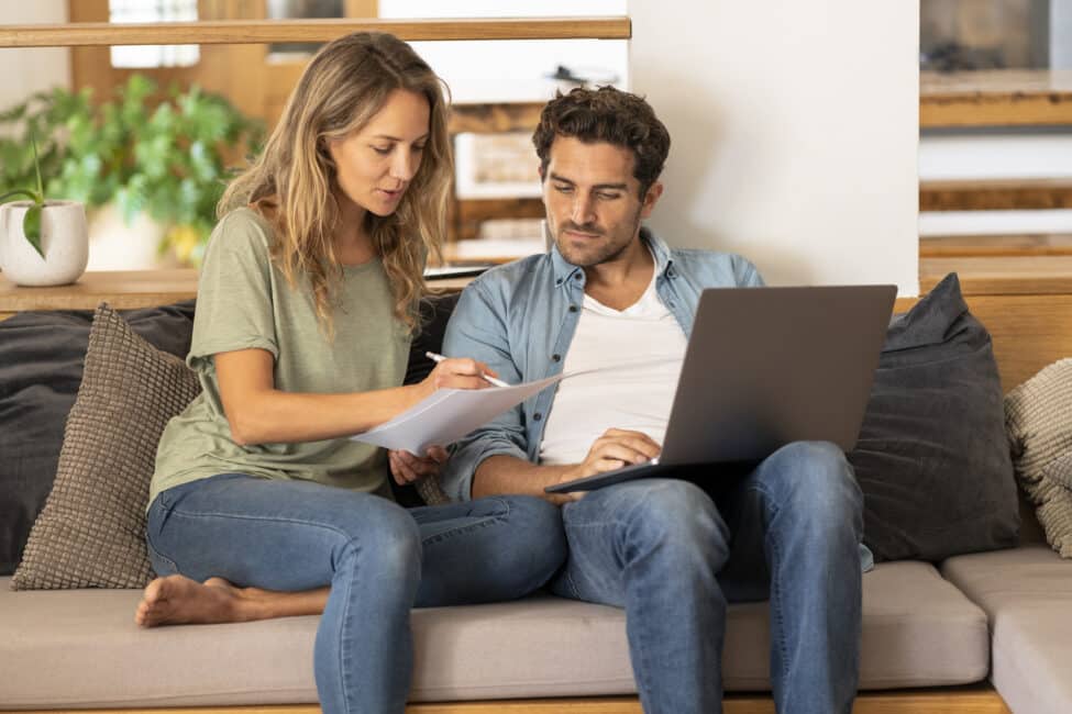 Concentrated couple at home sitting on couch in living room holding a laptop and signing papers