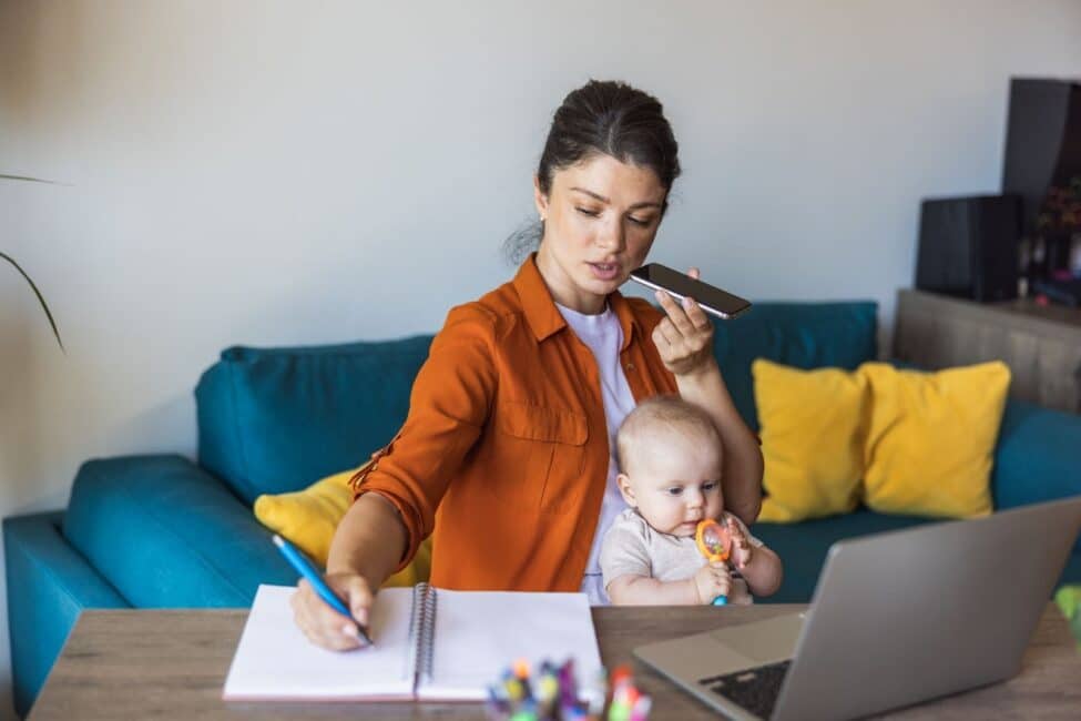 Busy mother with a baby on her lap, writing notes in a notebook while on the phone