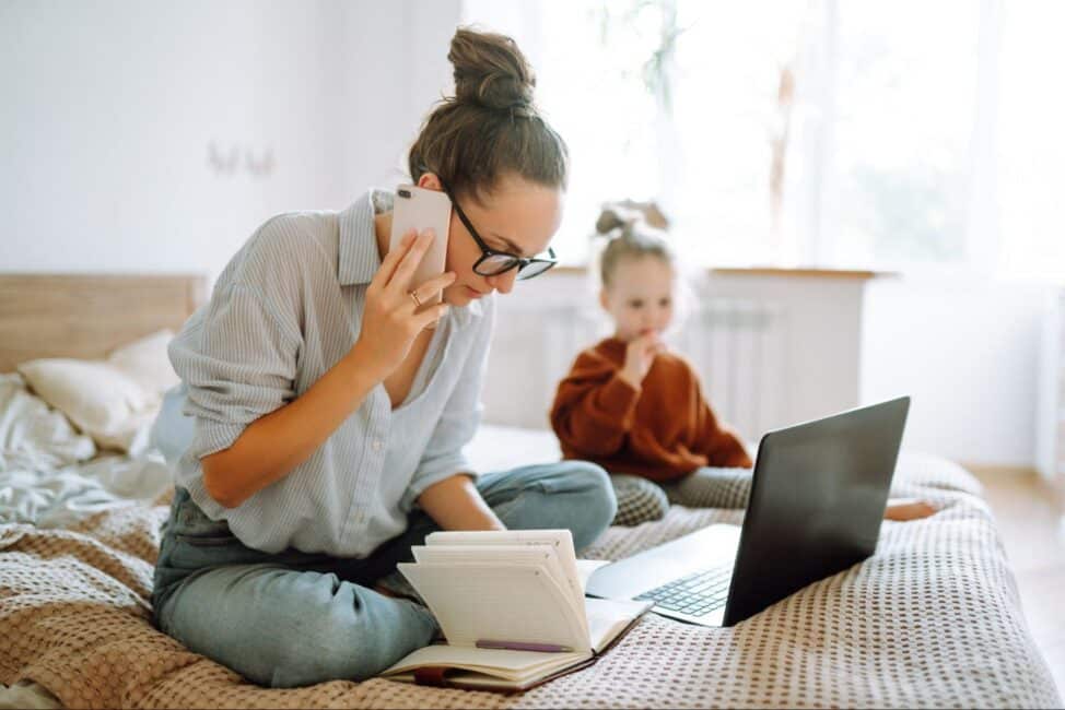 Focused mother talking on a smartphone while sitting cross-legged on a bed with her young daughter sitting quietly nearby