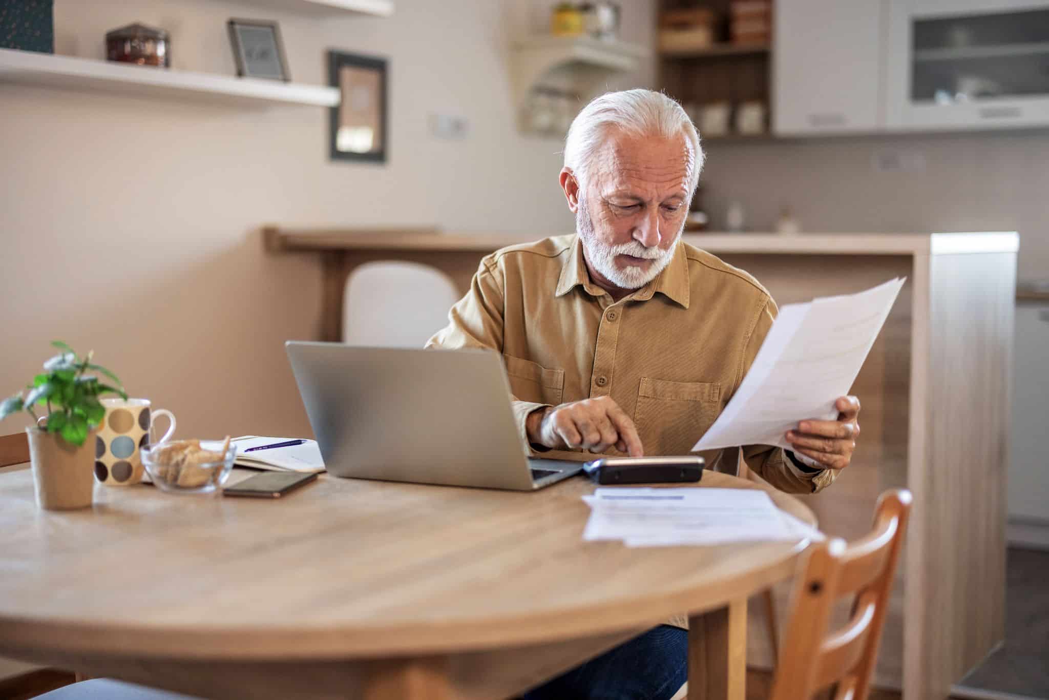 An elderly man reviewing financial documents and a balance transfer credit card offer on his laptop