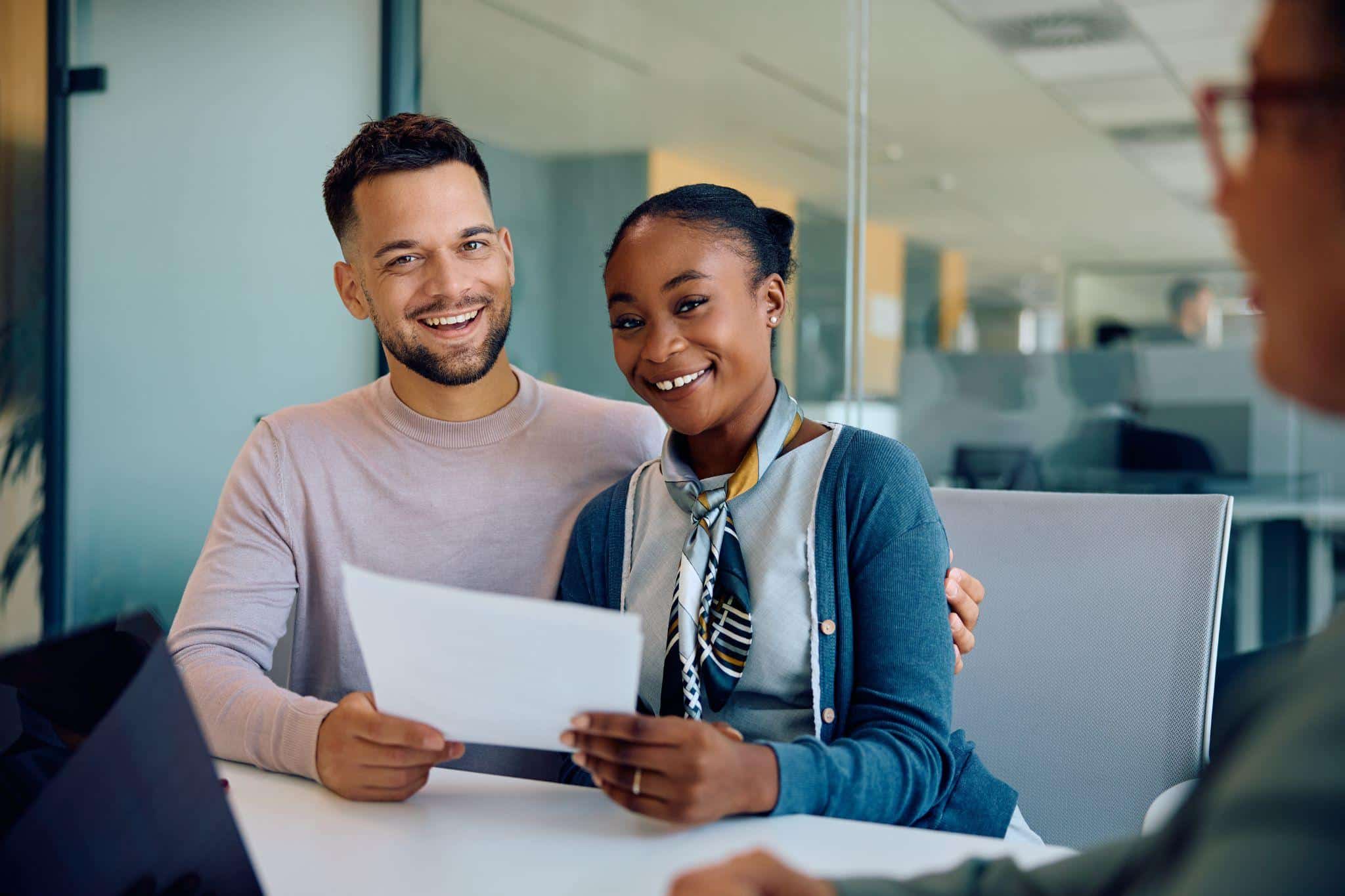 A smiling couple sits in a bright office, while meeting with a lender to increase their credit limit
