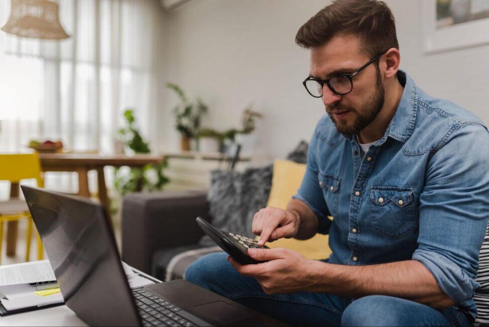 A focused man using a calculator and laptop to manage his finances and avoid overdraft fees