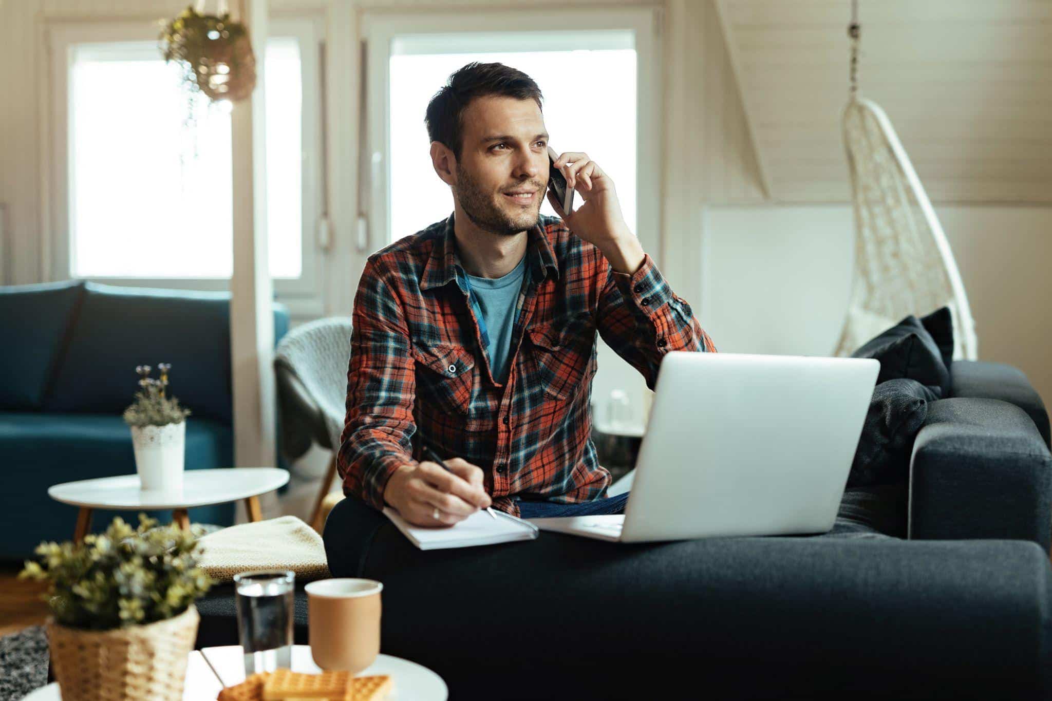Man talking on a phone while using a laptop and taking notes on a couch