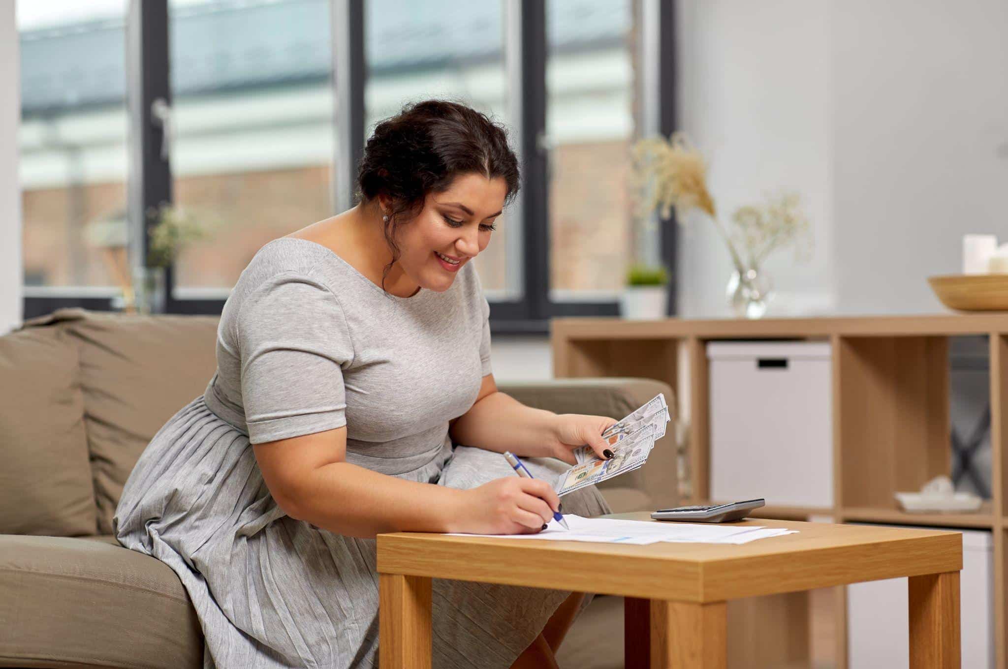 Smiling woman sitting on a couch, holding dollar bills while writing on financial papers next to a calculator