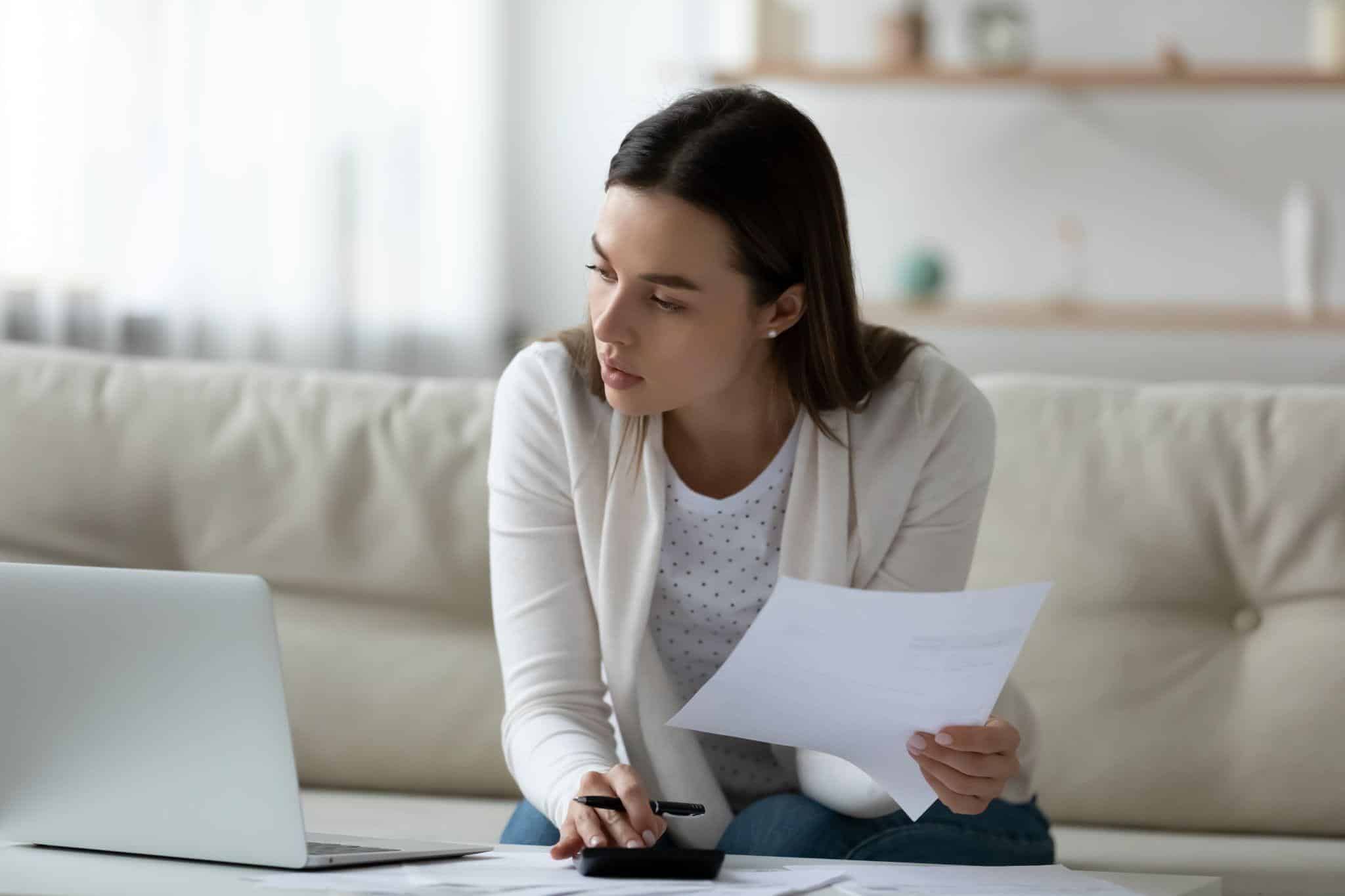 A woman looks at a laptop while holding papers and a pen and using a calculator to review her tax return