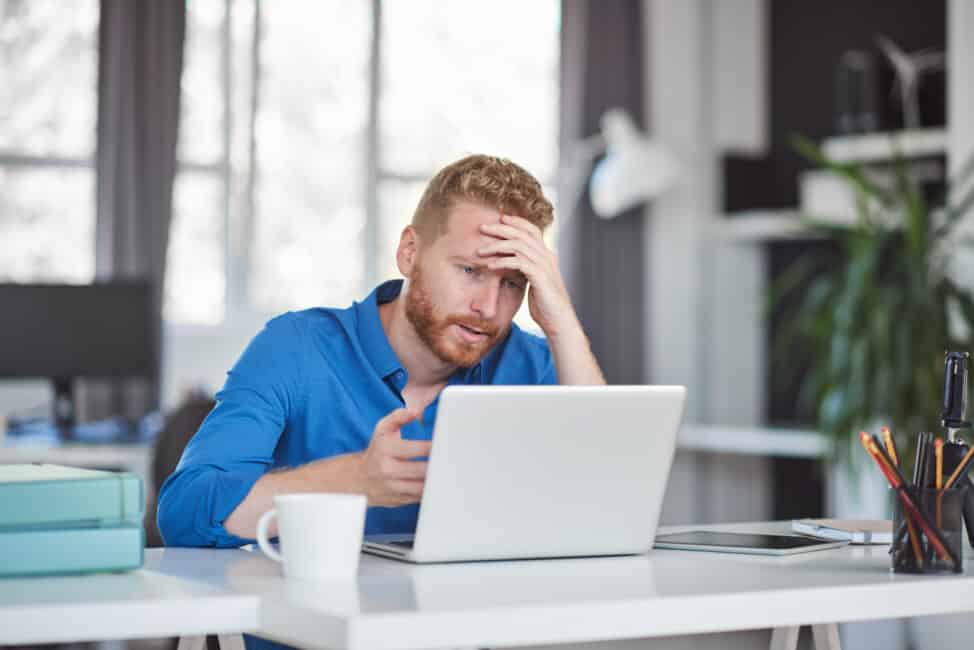 Stressed man in a blue shirt holding his forehead while looking at a laptop at his desk